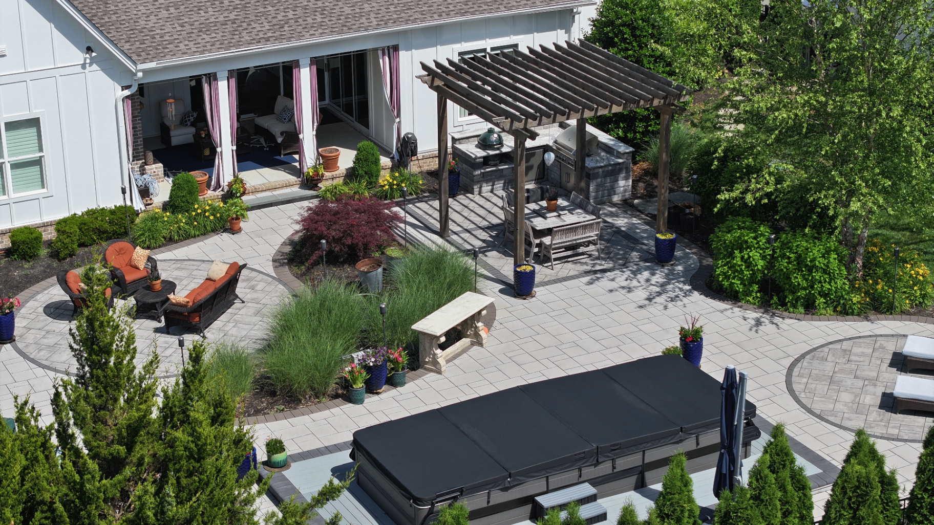 Wide aerial of paver patio with timber pergola, built-in grill island, covered porch, curved planting beds, and hot tub deck, Tennessee