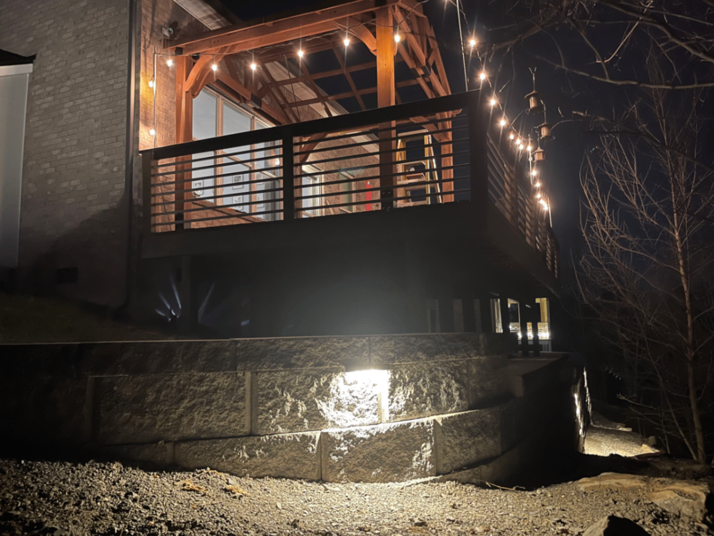 Timber-frame pergola over upper deck with string lights above lit stone retaining wall at night, Tennessee